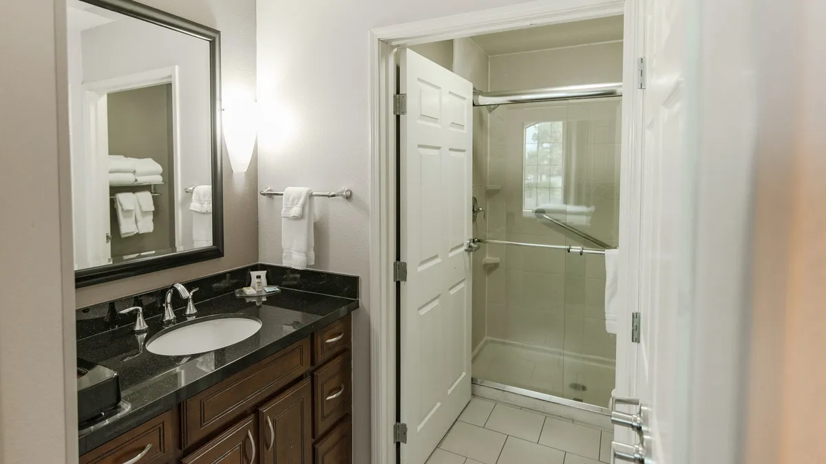 A stylish bathroom scene featuring a spacious vanity with a sink, a framed mirror, and a glimpse of the shower area. The space is well-lit, with modern fixtures that emphasize comfort and relaxation.