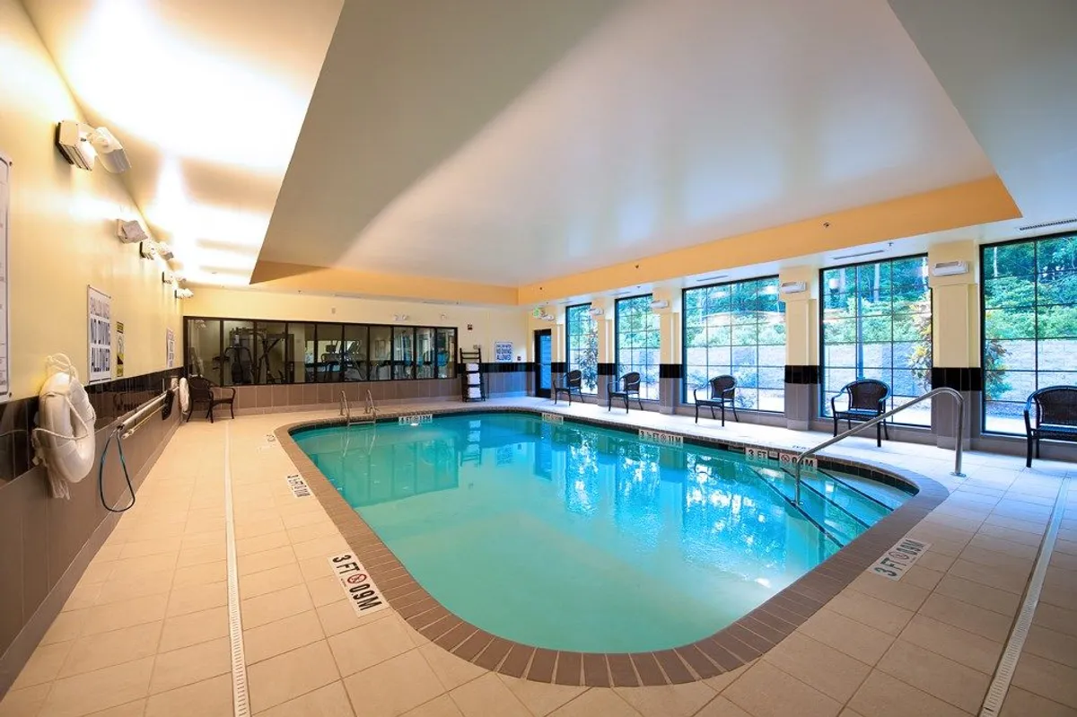 This image displays an inviting indoor swimming pool, bordered by large windows that allow natural light to fill the space. Comfortable chairs are arranged around the pool, providing spots for relaxation or socializing after a busy day. The setting suggests both tranquility and the opportunity for light activity.