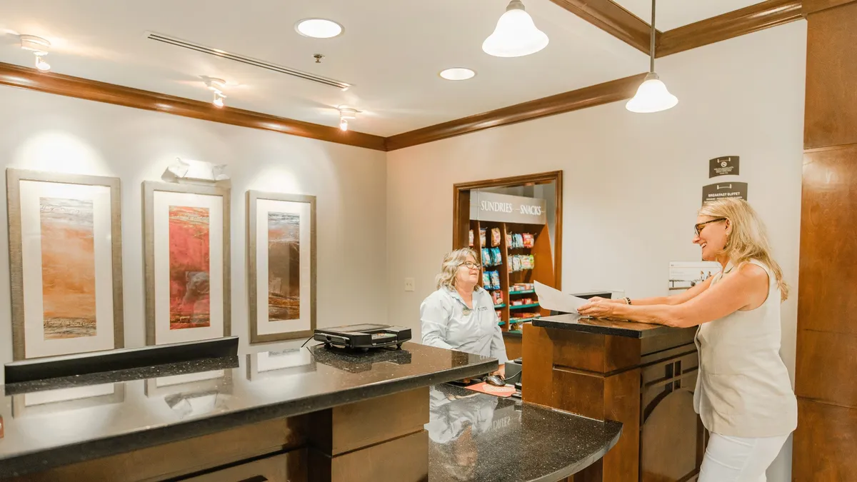 A welcoming reception area featuring a reception desk, two women engaged in a friendly check-in, contemporary art on the walls, and a convenient snack station. The scene exudes hospitality, guest service, and easy access to everyday needs.