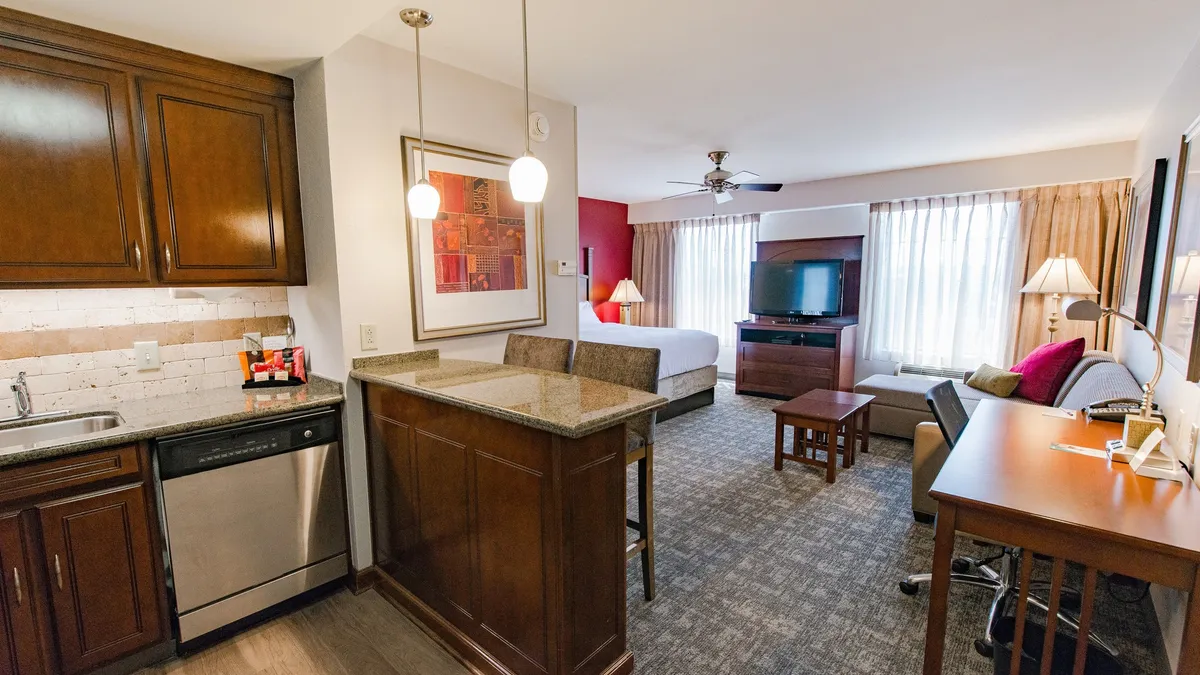 The image showcases a bright, modern studio with a well-equipped kitchen area, a cozy seating space perfect for relaxing or watching TV, and a plush king bed, all within an open, inviting layout. The overall atmosphere is both comfortable and functional, ideal for both leisure and productivity.