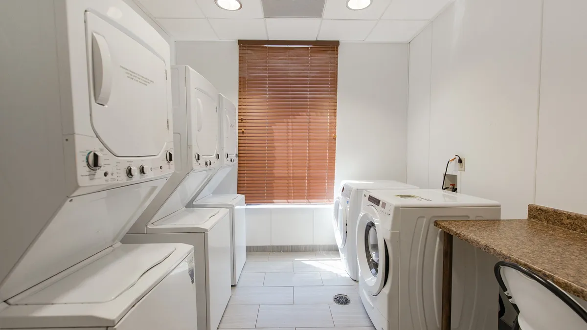 This image showcases a practical interior laundry area, featuring stacked washer and dryer units, a spacious countertop for sorting and folding clothes, and a window with blinds that brings in natural light. The space looks clean, organized, and efficient, perfectly suited for self-serve laundry tasks.